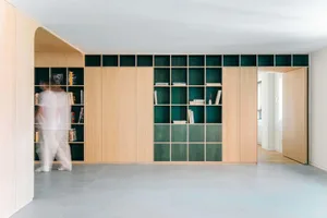 Modern minimalist living room with a large built-in light oak bookshelf and cabinet system featuring deep green painted back panels and open niches, grey tiled floor, and a person walking by.