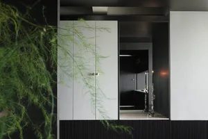 Modern dark entryway featuring white matte lacquer built-in cabinet, black ribbed paneling, two lit sculptural candlesticks, and a large hanging asparagus fern in the foreground.