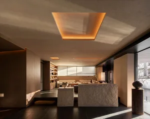 Modern minimalist living room featuring a large travertine central island, sunken seating area, built-in wooden shelving, and a glowing rectangular skylight casting golden light across the dark tiled floor.