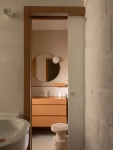 View into a modern bathroom with a peach-colored vanity, round mirror, white wall sconce, and a speckled terrazzo stool in the foreground.