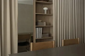 Modern minimalist dining area featuring light oak built-in bookshelves, wooden dining chairs, neutral beige curtains, and decorative objects on display.