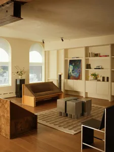 Modern living room featuring a raised burl wood platform, a corduroy-upholstered wooden sofa, four concrete cube stools on a striped rug, built-in shelving, and arched windows.