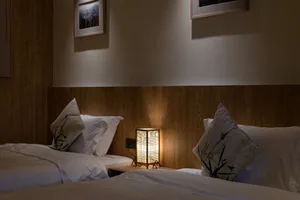 Cozy twin beds in a Japanese-inspired bedroom featuring vertical wood paneling, white bedding with bamboo-print pillows, and a lit rectangular lantern on a wooden nightstand.