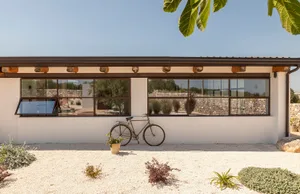 Modern white stucco farmhouse exterior with large dark-framed windows, exposed wooden beams, a vintage bicycle leaning against the wall, and a gravel courtyard with native plants under a clear blue sky.