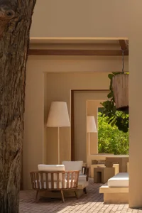 Wooden teak swivel armchair with white cushions placed on a brick-paved courtyard next to concrete outdoor furniture and large floor lamps, framed by a textured tree trunk and lush green foliage in the background.