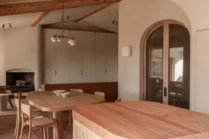 Modern minimalist dining room featuring a large oval oak dining table, wooden chairs, a terracotta stone kitchen island in the foreground, built-in banquette seating, exposed wooden ceiling beams, and a fireplace in the background.
