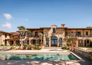 Front view of a luxurious Mediterranean-style stone villa with a rectangular swimming pool in the foreground, arched windows, terracotta roof tiles, and lush tropical landscaping under a clear blue sky.
