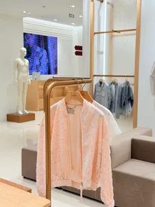 A soft pink fuzzy bomber jacket and a white top hanging on a brass clothing rack in a modern minimalist retail store with beige seating and a mannequin in the background.
