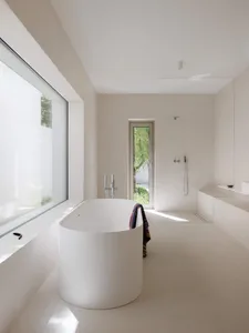 Minimalist bathroom interior featuring a white oval freestanding bathtub, large picture window, concrete walls and floor, and a glass-enclosed shower area with natural daylight.
