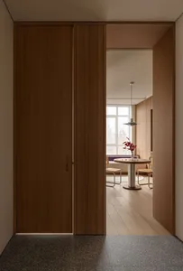 Modern minimalist interior showing a tall walnut wood pivot door partially open, revealing a bright dining area with round wooden table, woven chairs, pendant light, and window seat in the background.