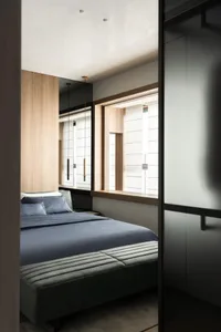Modern minimalist bedroom with blue bedding, grey upholstered bench, light wood wall paneling, and long pendant lights hanging beside a large window.