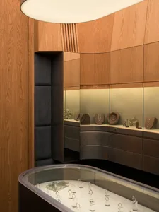 Interior view of a luxury jewelry store featuring curved walnut wood cabinetry, dark padded wall panels, and illuminated glass display cases with gold jewelry.
