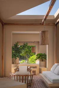 Open-air courtyard patio featuring wooden ceiling beams, a cylindrical wooden pendant lamp, teak armchair with white cushions, built-in stone sofa, and lush potted plants under a bright blue sky.