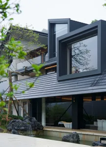 Modern black steel villa exterior with large picture windows, dark tiled roof, reflecting pool, large rocks, and a small waterfall surrounded by lush greenery.