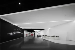 Modern minimalist exhibition hall with black and white walls, glossy dark floor, and multiple futuristic concept cars displayed on white pedestals, featuring a large black wall with a glowing white car silhouette.