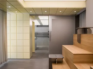 Modern minimalist entryway showing light oak wooden stairs, a built-in bench, pale yellow perforated wall panels, concrete floor, and a view into a kitchen area with stainless steel elements.