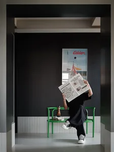 Person reading a newspaper while sitting on a bright green chair in a modern dark entryway with a large Alexander Calder poster on the wall.