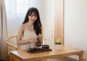 Woman with long dark hair wearing a beige dress, seated at a light wooden table performing a Japanese tea ceremony with a black ceramic teapot and cups, next to a small white vase with moss and flowers.