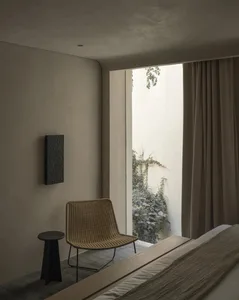 Minimalist bedroom interior featuring a woven rattan chair, dark side table, textured wall art, and large opening to a lush green courtyard with beige curtains.
