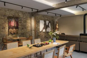 Modern industrial dining room with exposed stone walls, long light oak dining table, wooden rattan chairs, brown leather sofa, and contemporary art pieces.