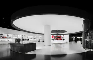 Interior view of a modern automotive museum exhibition hall featuring a large circular white ceiling structure, a suspended red Ferrari car chassis, glossy black floor, and illuminated display cases along the curved walls.