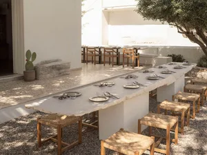 Long outdoor dining table made from natural travertine stone with woven rush stools arranged on gravel in a sunny white-walled Mediterranean courtyard with a prickly pear cactus.