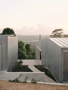 Modern house clad in weathered Corten steel with wooden deck and stairs, set against a vast forested mountain valley at dusk.