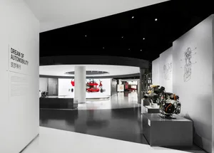 Modern exhibition hall displaying automotive engines, technical diagrams on walls, and a suspended red car in the background under dramatic lighting.