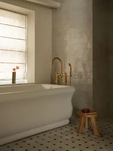 A serene bathroom corner featuring a white freestanding bathtub, unlacquered brass faucet, wooden stool, and beige zellige tiled walls with a window dressed in natural linen roman shades.