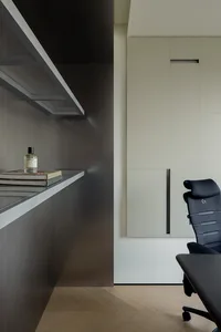 Modern minimalist home office corner featuring a matte black built-in wall unit, floating shelf with books and perfume, light beige flush cabinetry, and a black ergonomic office chair on light wood herringbone flooring.
