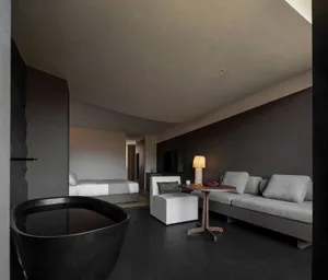 Modern minimalist bedroom and living area featuring a prominent black freestanding stone bathtub in the foreground, light grey modular sofa, round wooden side table with lamp, and curved micro-cement walls and ceiling.