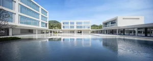 Modern minimalist building complex with a large rectangular reflection pool in the courtyard, featuring white concrete structures and extensive glass windows under a clear blue sky.