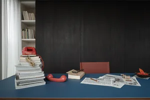 Blue desk with stacks of books, a red vintage rotary telephone, scattered newspapers, a magnifying glass, and a red leather chair in front of dark wood paneling and a built-in bookshelf.