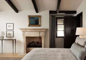 Cozy bedroom interior with a large bed with neutral linen bedding, an antique carved limestone fireplace with brick interior, dark wooden ceiling beams, and framed artwork on white walls.