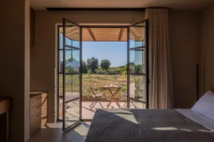 Modern minimalist bedroom featuring a large bed with grey linen, light wood dresser, and wide open black-framed bifold glass doors revealing a vineyard terrace and rural landscape beyond.