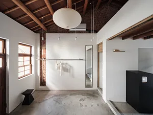Bright industrial-style entryway with white walls, exposed red brick, polished concrete floor, wooden ceiling beams, large white paper lantern, black console table, and full-length mirror.