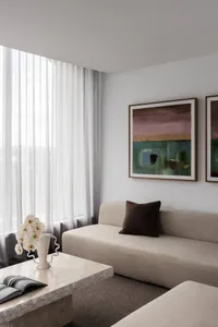 Modern minimalist living room featuring a beige sofa with brown cushion, a large travertine coffee table with white orchid arrangement, and two abstract landscape paintings on a white wall.