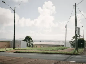 一座低矮的工業建築，擁有波紋金屬屋頂和混凝土牆，坐落在鄉村山坡上，周圍環繞著電線桿和綠草，頭頂是明亮多雲的天空。