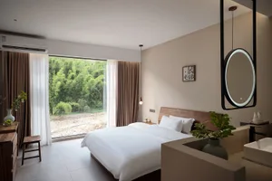 Modern hotel bedroom featuring a walnut wood headboard, white bedding, large glass doors opening to a bamboo forest, pendant light, and integrated vanity with round backlit mirror.
