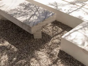 Close-up view of a thick travertine stone bench resting on a bed of grey gravel, with strong tree shadows cast by bright sunlight.