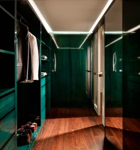 Narrow walk-in closet featuring glossy emerald green lacquered cabinetry, open shelving with hanging clothes and shoes, rich walnut hardwood floors, and continuous linear LED cove lighting along the ceiling.