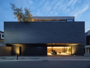 Front view of a modern two-story house with dark charred wood cladding, a large open carport containing a black sedan and motorcycle, and rooftop greenery against a blue dusk sky.