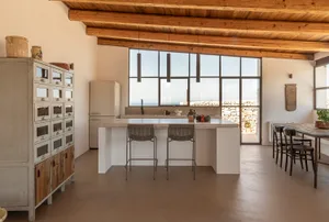 Modern rustic open-plan kitchen featuring a large terrazzo island with woven bar stools, exposed wooden ceiling beams, white cabinetry, and large windows overlooking the sea and stone terrace.
