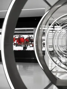 View through large circular steel and glass frames into a modern exhibition hall displaying red car chassis and mechanical components on a white platform.