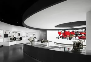 Interior view of a modern automotive museum with suspended red cutaway car sections, large mechanical components on glossy black pedestals, and illuminated display cases along curved white walls.