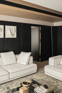 Modern living room featuring a black oak accent wall, white sofas with black piping, abstract wall art, and a patterned area rug with a round glass coffee table.