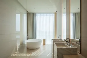 Modern minimalist bathroom featuring a white freestanding oval bathtub, double marble rectangular sinks with chrome faucets, large mirrors, sheer curtains, and abundant natural light.