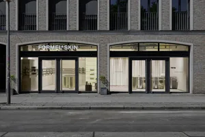 柏林 Formel Skin 美容診所店面街景，兩個拱形玻璃入口搭配黑色框架、黃銅圓柱展示桌、淡黃色內牆，以及人行道上的盆栽樹木。