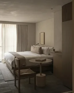 Minimalist bedroom interior featuring a low wooden platform bed with white and beige linen bedding, a woven rope chair, round concrete side table, and warm oak wood wall paneling under soft daylight.