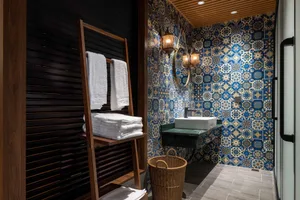 Compact powder room featuring vibrant blue and gold Moroccan patterned tiles, white vessel sink on green marble countertop, round mirror with wall sconces, wooden ladder towel rack with white towels, and woven basket on dark grey floor tiles.